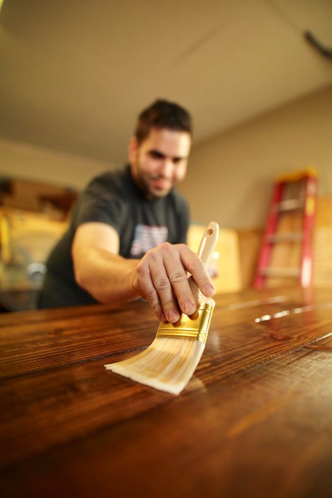 pexels-photo-3186683 A man carefully applies varnish to a wooden surface indoors, using a paintbrush for a smooth finish.