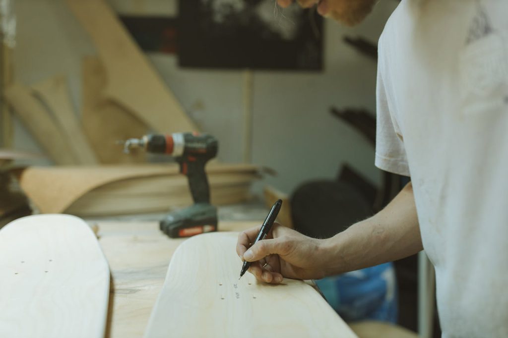 pexels-photo-4889069-1 A craftsman in a workshop marks a skateboard deck with precision tools, surrounded by wood and drill.